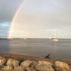 A rainbow arcs down to the breakwater and the lighthouse in Manitowoc, WI. A boat is visible on the water.